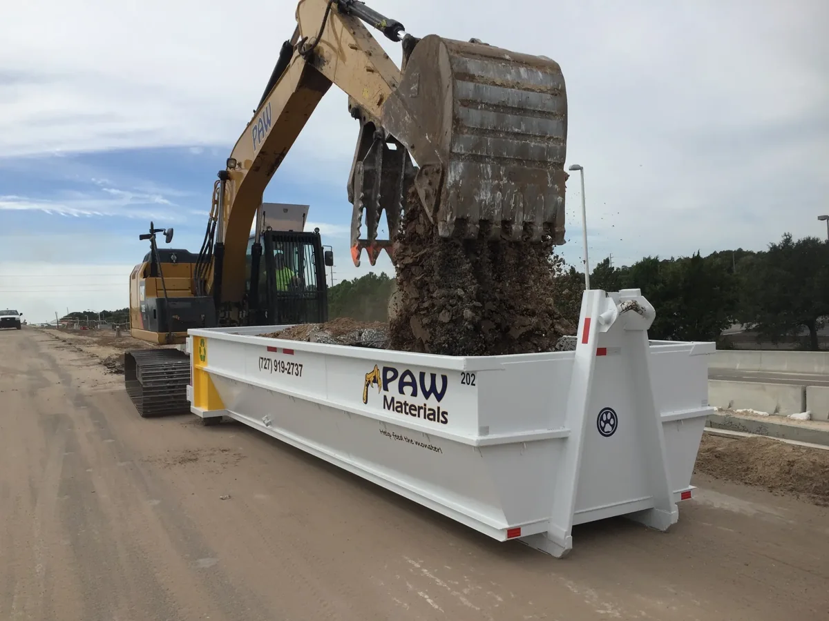 Excavator loading a PAW Materials roll-off dumpster on a commercial job site